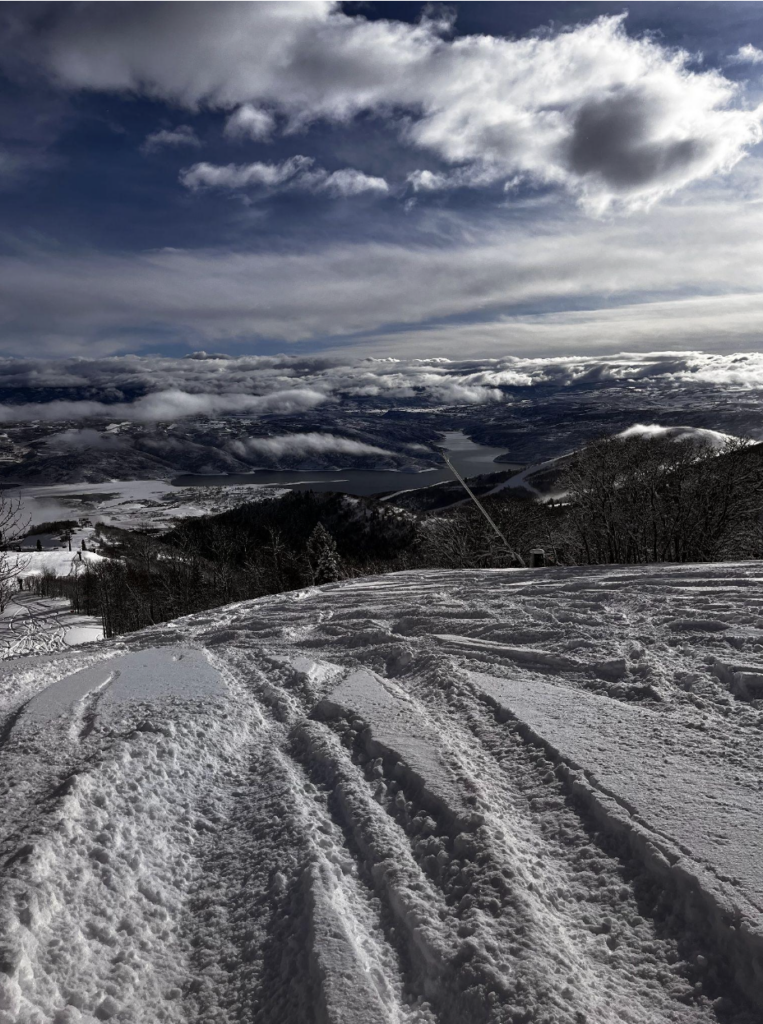 Snowy mountain slope with fresh ski and snowboard tracks, overlooking a valley with a lake and scattered clouds—a perfect scene for any ski and snowboard club adventure.
